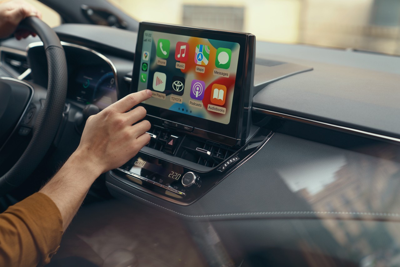 O persoana din interiorul unei Toyota interactioneaza cu ecranul tactil multimedia al masinii. Afisajul arata ecranul de pornire Apple CarPlay.